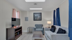 Ein modernes Wohnzimmer mit Flachbildfernseher, einem grauen Sessel, einem grauen Sofa, einem Couchtisch und einem Beistelltisch mit Lampe.