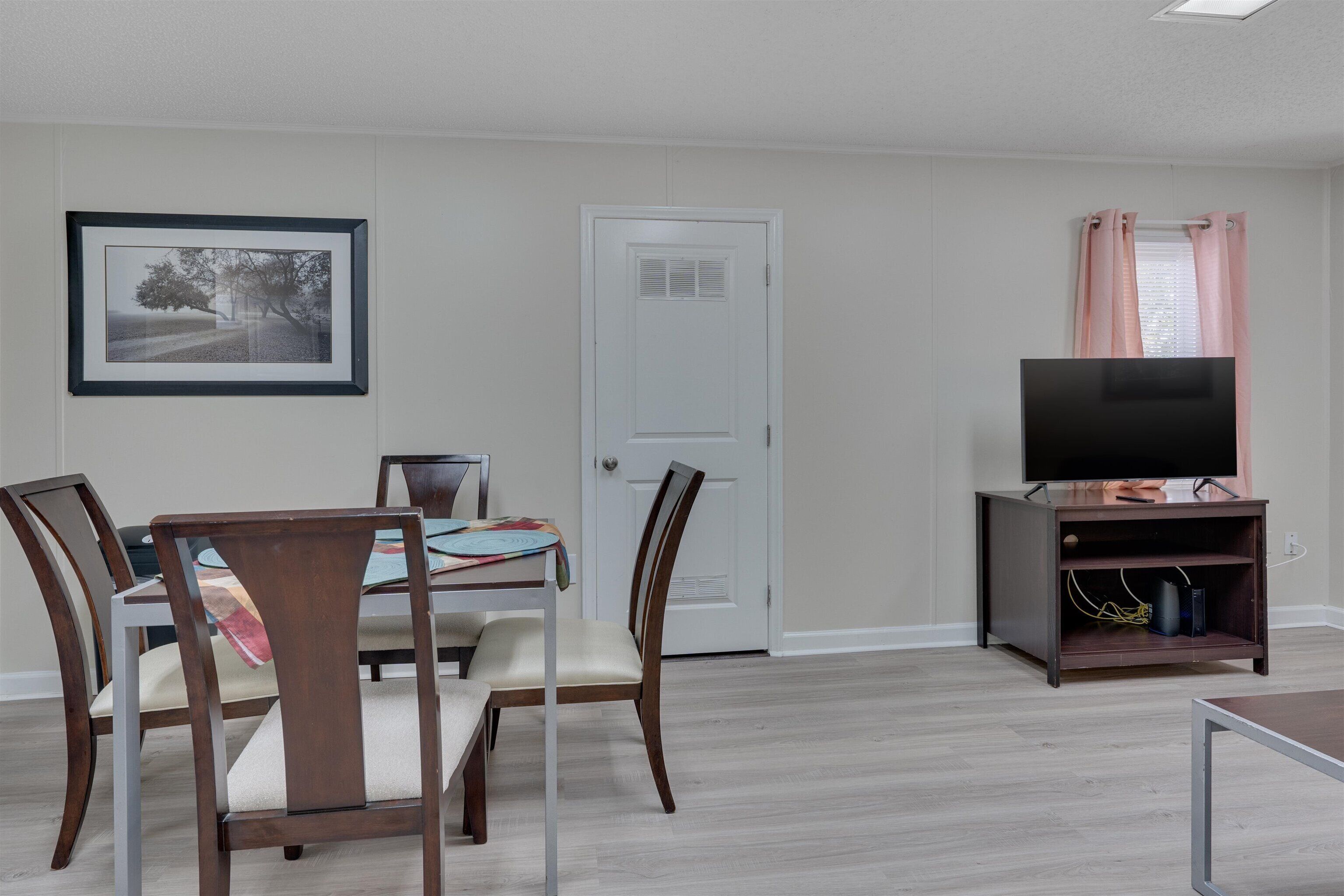 Un coin repas avec une table et des chaises, un tableau encadré au mur, une porte fermée et un téléviseur posé sur un meuble.