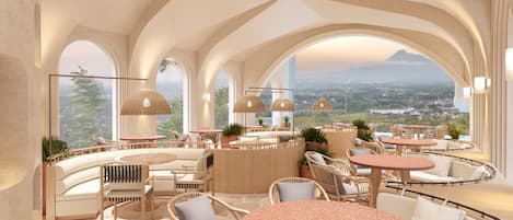 A spacious lounge area with round tables and wicker chairs, large arched windows offering a view of the landscape, and pendant lighting.