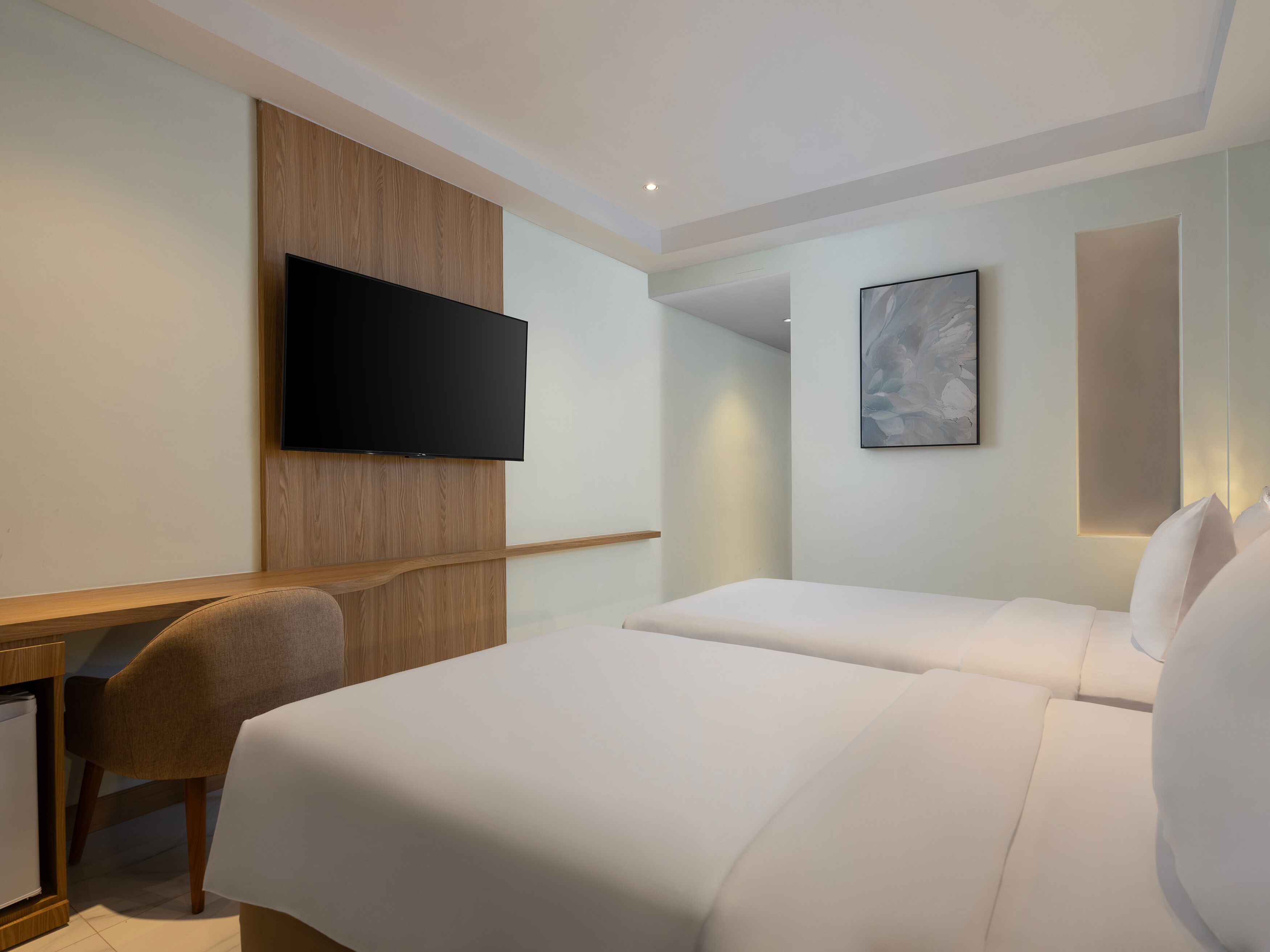 A hotel room with a flat-screen TV mounted on a wooden panel, a desk with a chair, two beds with white bedding, and a framed abstract painting on the wall.