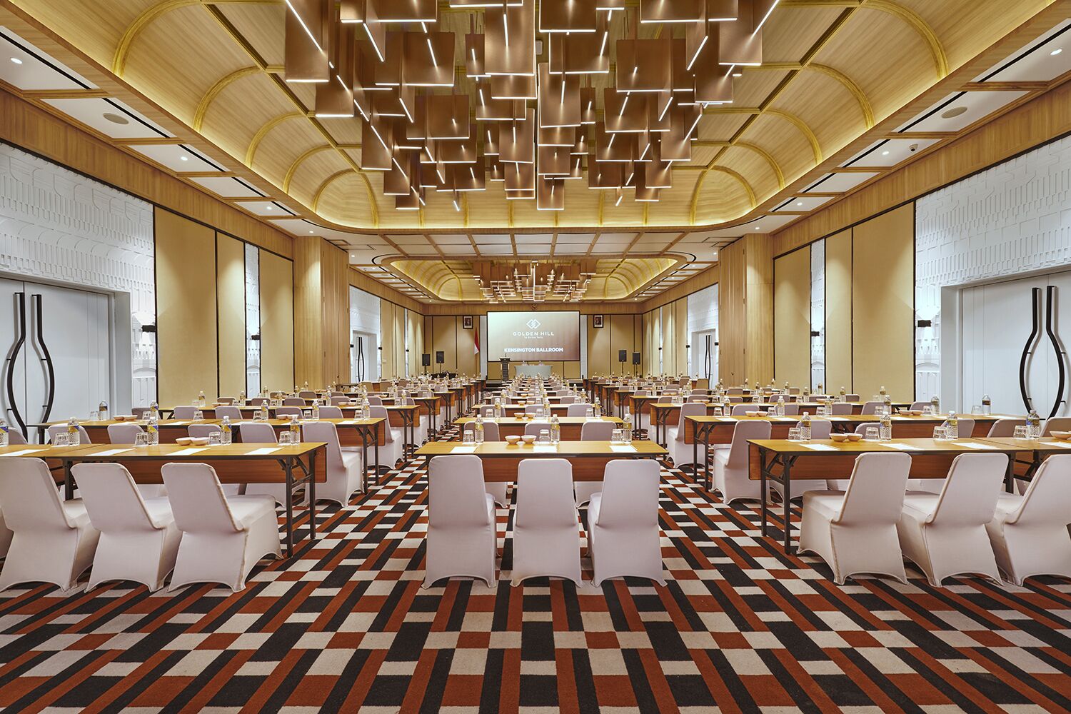 Un amplio salón de conferencias con filas de mesas y sillas, una alfombra estampada y un diseño moderno en el techo.