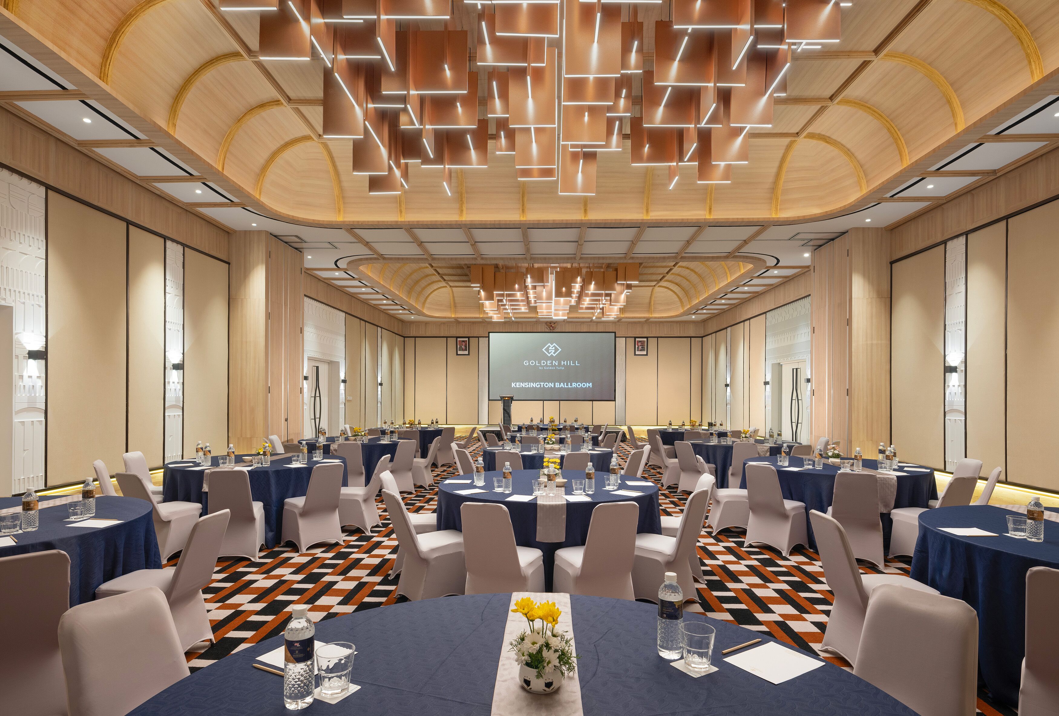 Ein Konferenzraum mit runden Tischen, Stühlen und einem großen Bildschirm, der "GOLDEN HILL KENSINGTON BALLROOM" anzeigt.