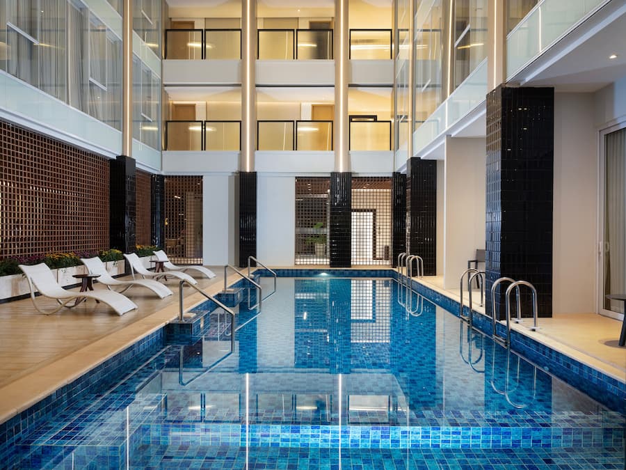 Una piscina interior con baldosas azules brillantes, rodeada de sillas de descanso y una barandilla de cristal.