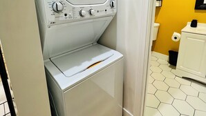 A compact, white washer and dryer unit in a small laundry room with a yellow wall and a hexagonal tile floor.