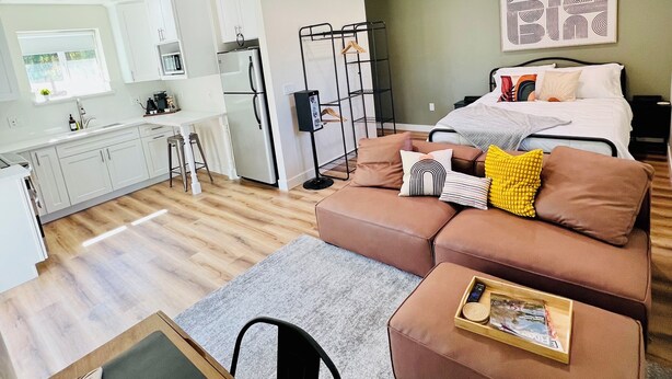 A modern apartment with a kitchenette, a living area with a sofa and ottoman, a dining table, and a ceiling fan.