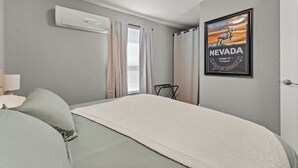 A hotel room with a bed, a nightstand, a lamp, a poster on the wall, and an air conditioning unit. - Tranquil Retreat: Cozy 2Bed, 1Bath Oasis in Fallon (Fallon)