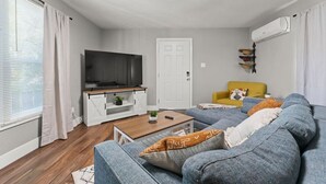 A modern living room with a large grey sofa, a wooden coffee table, a flat-screen TV on a white cabinet, a yellow armchair, and a white door. - Tranquil Retreat: Cozy 2Bed, 1Bath Oasis in Fallon (Fallon)