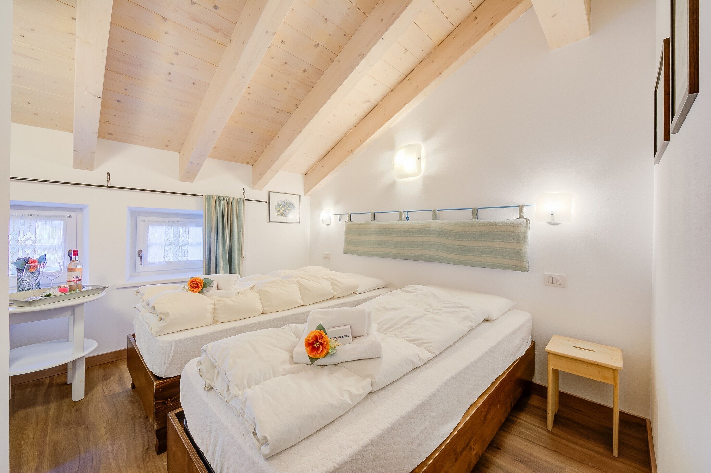 A room with two beds, a small table, a wall-mounted light, and a window with curtains.