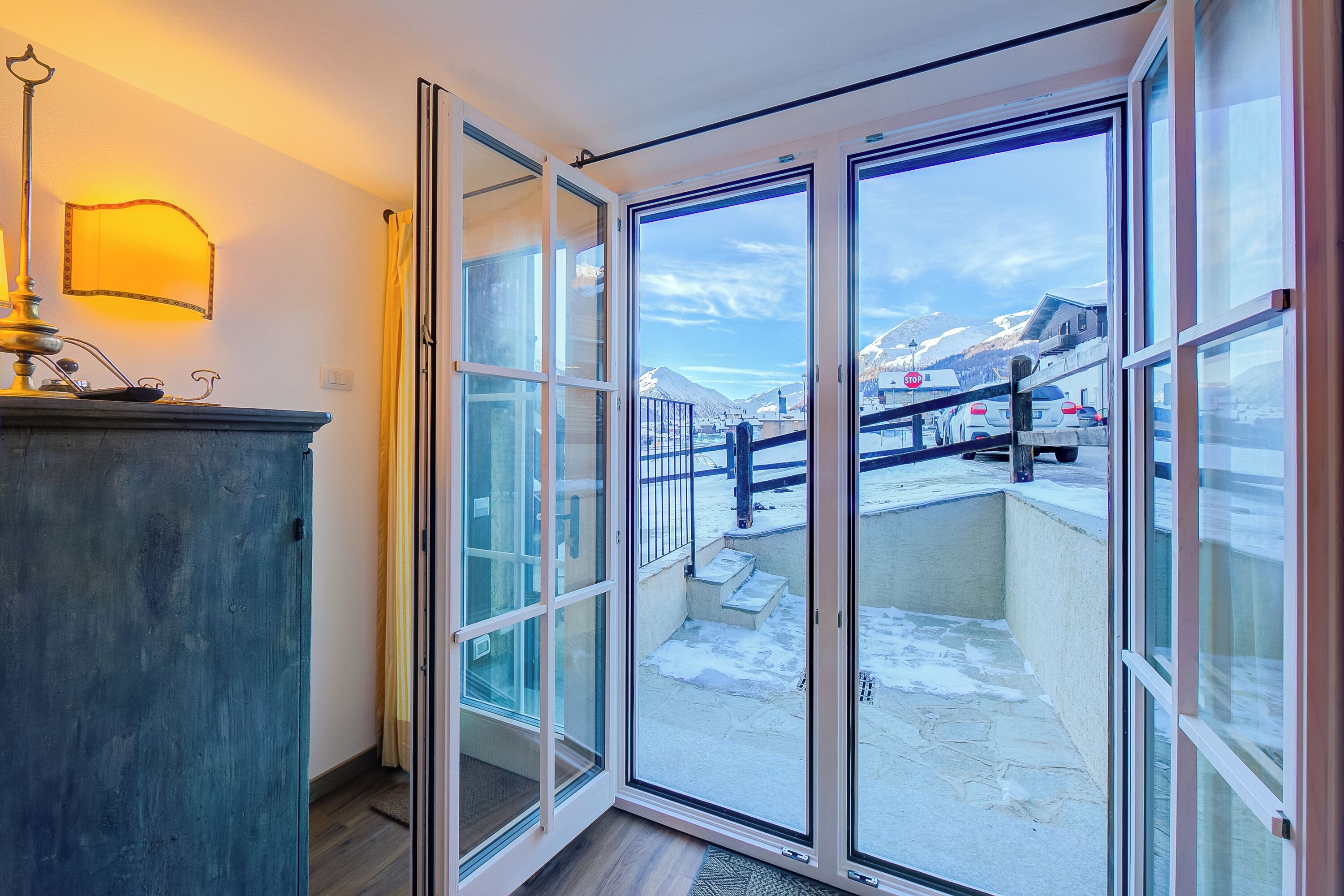 Una habitación con una puerta corrediza de vidrio que da a un balcón con vistas a una montaña nevada y coches aparcados.