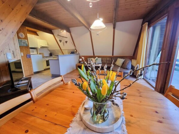 Un intérieur chaleureux, avec des éléments en bois, comprenant un coin repas doté d’une table agrémentée d’un vase de fleurs. On aperçoit une cuisine au fond.