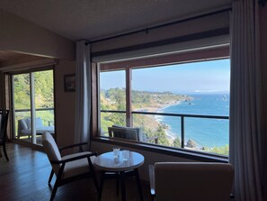 A room with a large window overlooking a coastal landscape, a round table with two chairs, and a white sofa.
