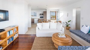 A modern living room with a white sofa, a wooden bookshelf, a round coffee table, and a patterned sofa. - Beachies - Oceanstays (Angourie)