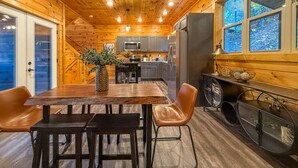 A wooden cabin interior with a dining area, kitchen, and large windows.