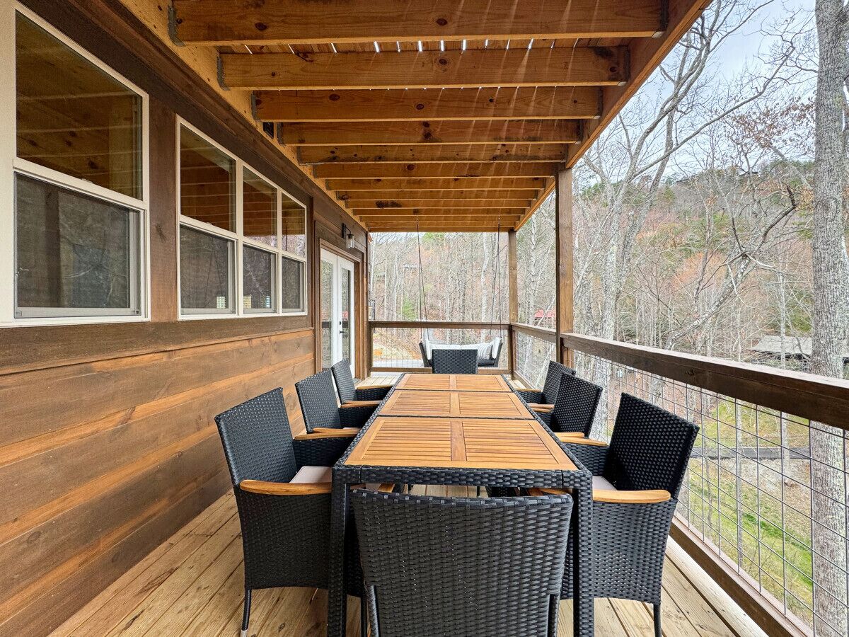 A covered outdoor area with a wooden deck, a long table with chairs, and a view of trees and buildings in the background.