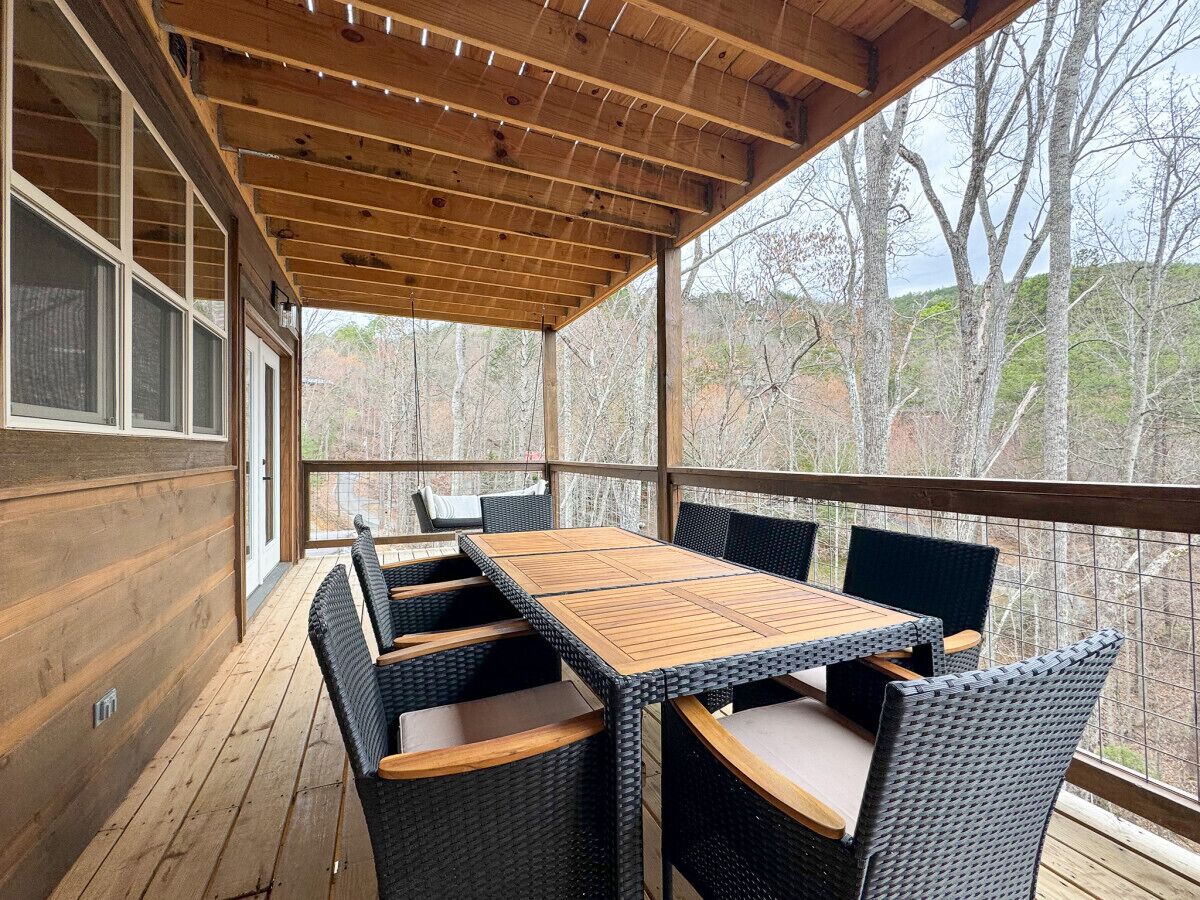 A covered outdoor seating area with a wooden table and wicker chairs, overlooking a forested area.