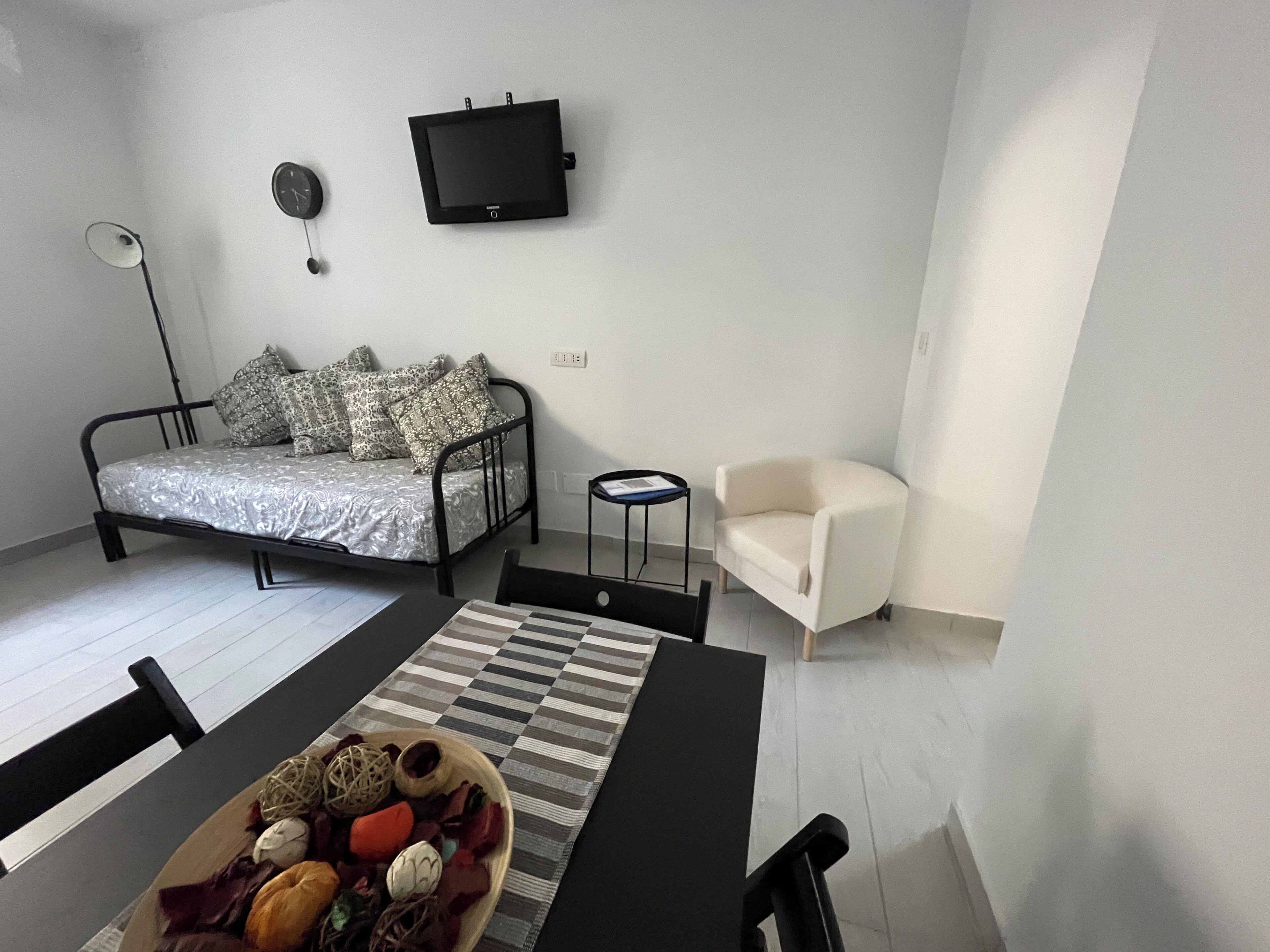 Una stanza moderna e accogliente con un letto, una televisione a parete, un piccolo tavolo con una ciotola di frutta, una sedia e una lampada.