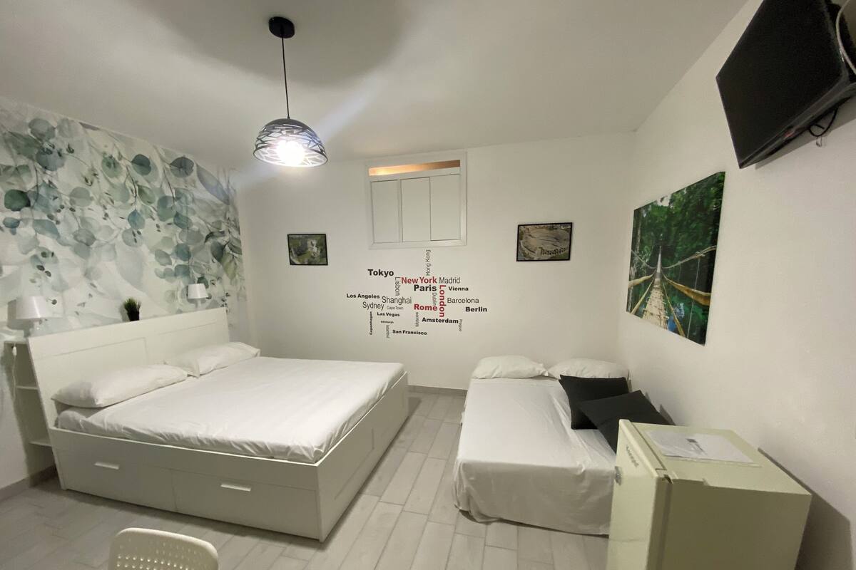 A bedroom with a bed, bedside tables, a chair, a wardrobe, a wall with city names, a window, and a hanging light fixture.