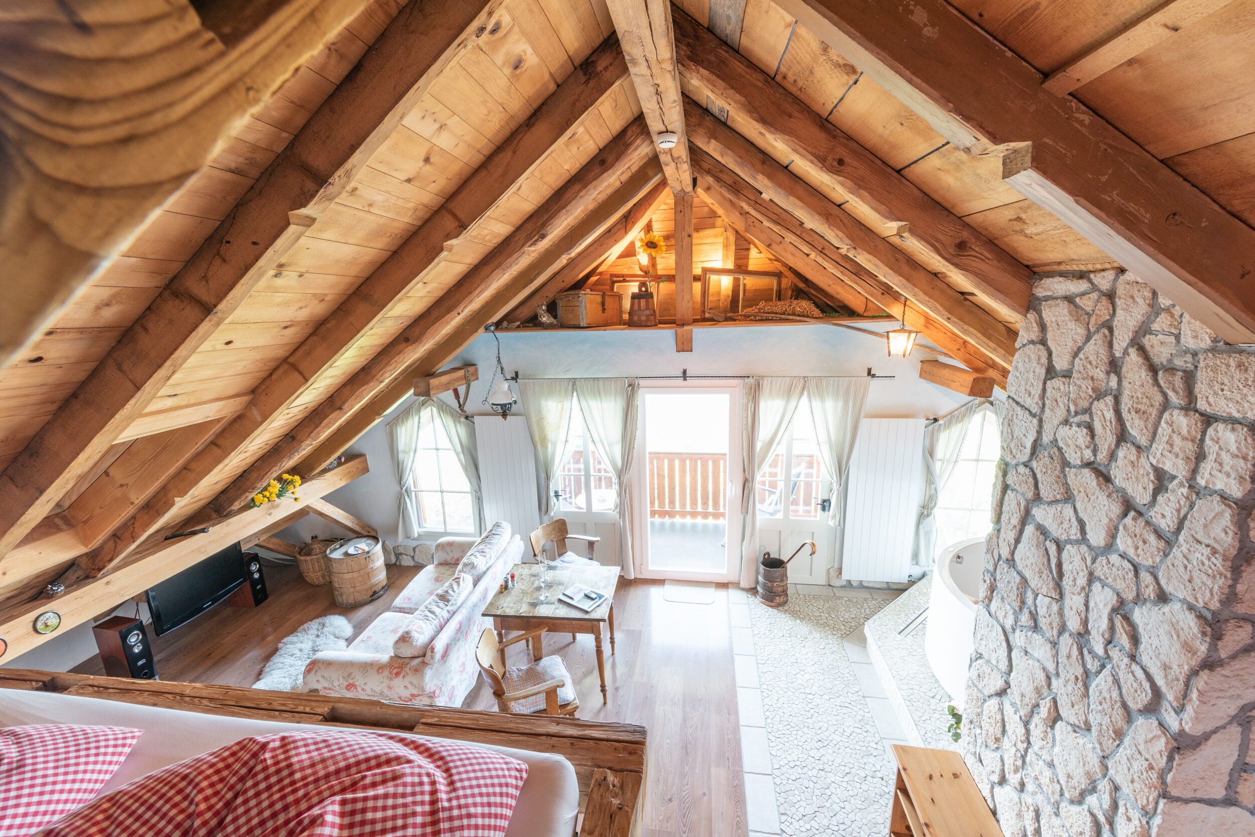 An attic bedroom with a stone wall, wooden beams, a bed, a table, and a sofa.