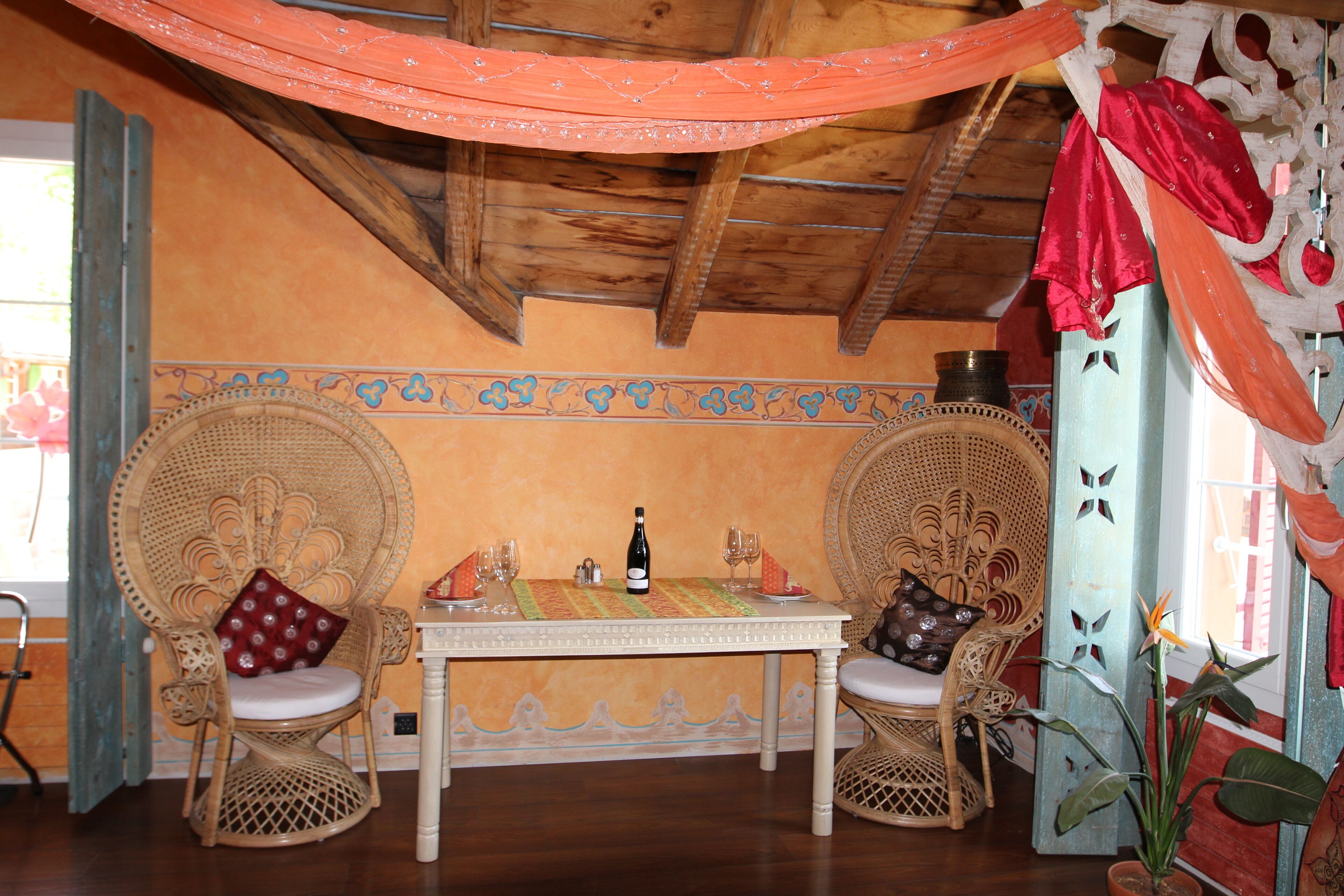 A room with a wooden ceiling, two wicker chairs with cushions, a table with a bottle and glasses, and a decorative wall with a floral pattern.