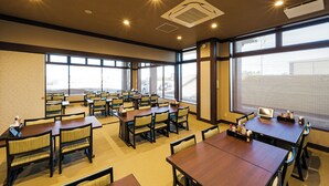 A hotel dining area with multiple tables and chairs, large windows, and ceiling lights.