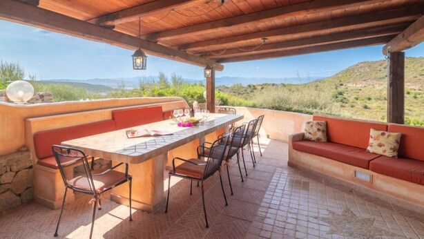 An outdoor patio with a wooden ceiling, a stone wall, and a view of a hilly landscape. - Vacation home Fusco by Interhome (Olbia)