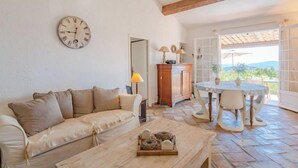 A cozy living room with a beige sofa, a wooden coffee table, and a dining table with chairs. A wooden cabinet and a wall clock are also visible. - Le Clos d´Estelle by Interhome (La Garde Freinet)