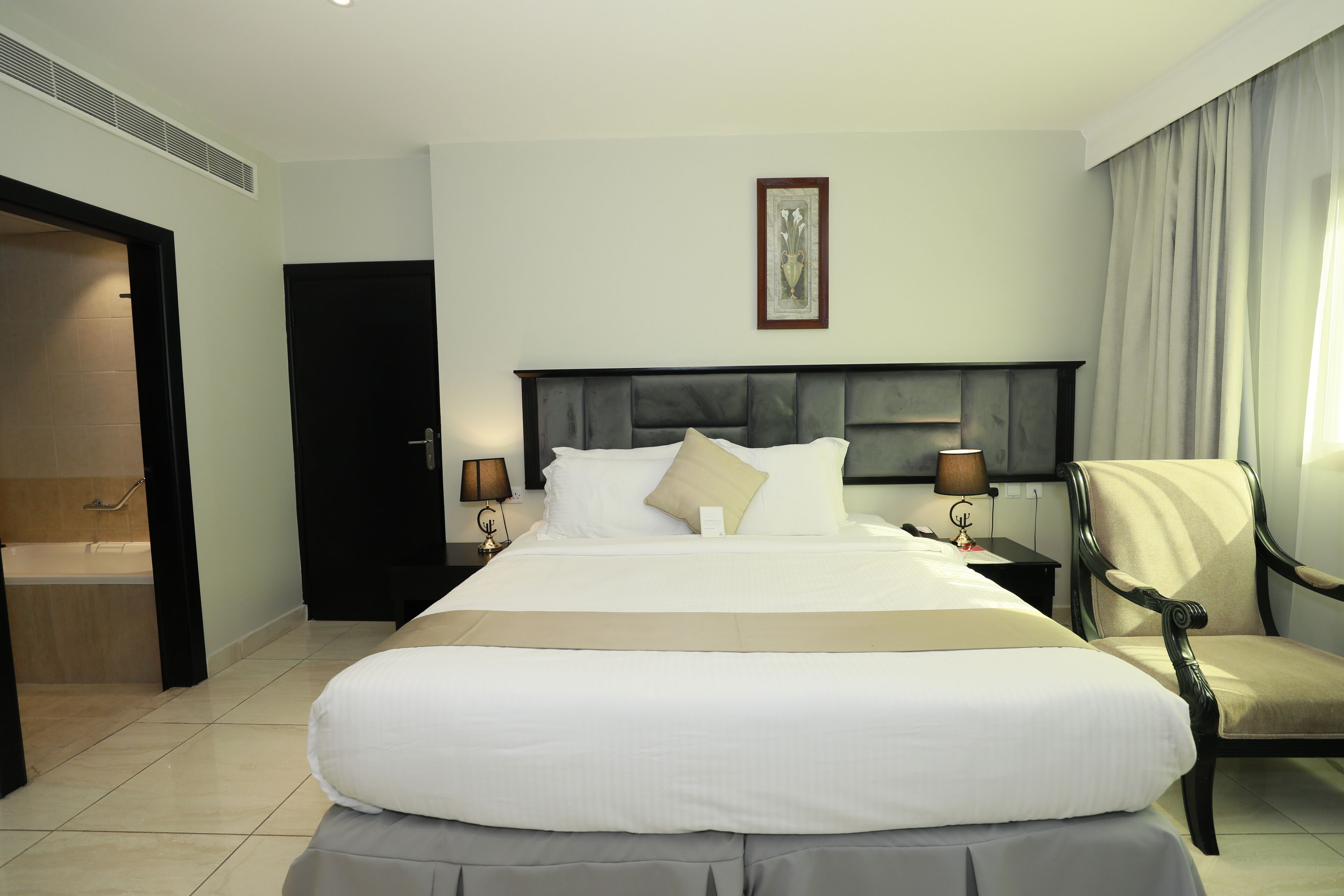 Una habitación de hotel con una cama grande, lámparas de noche, una silla y un cuadro en la pared.