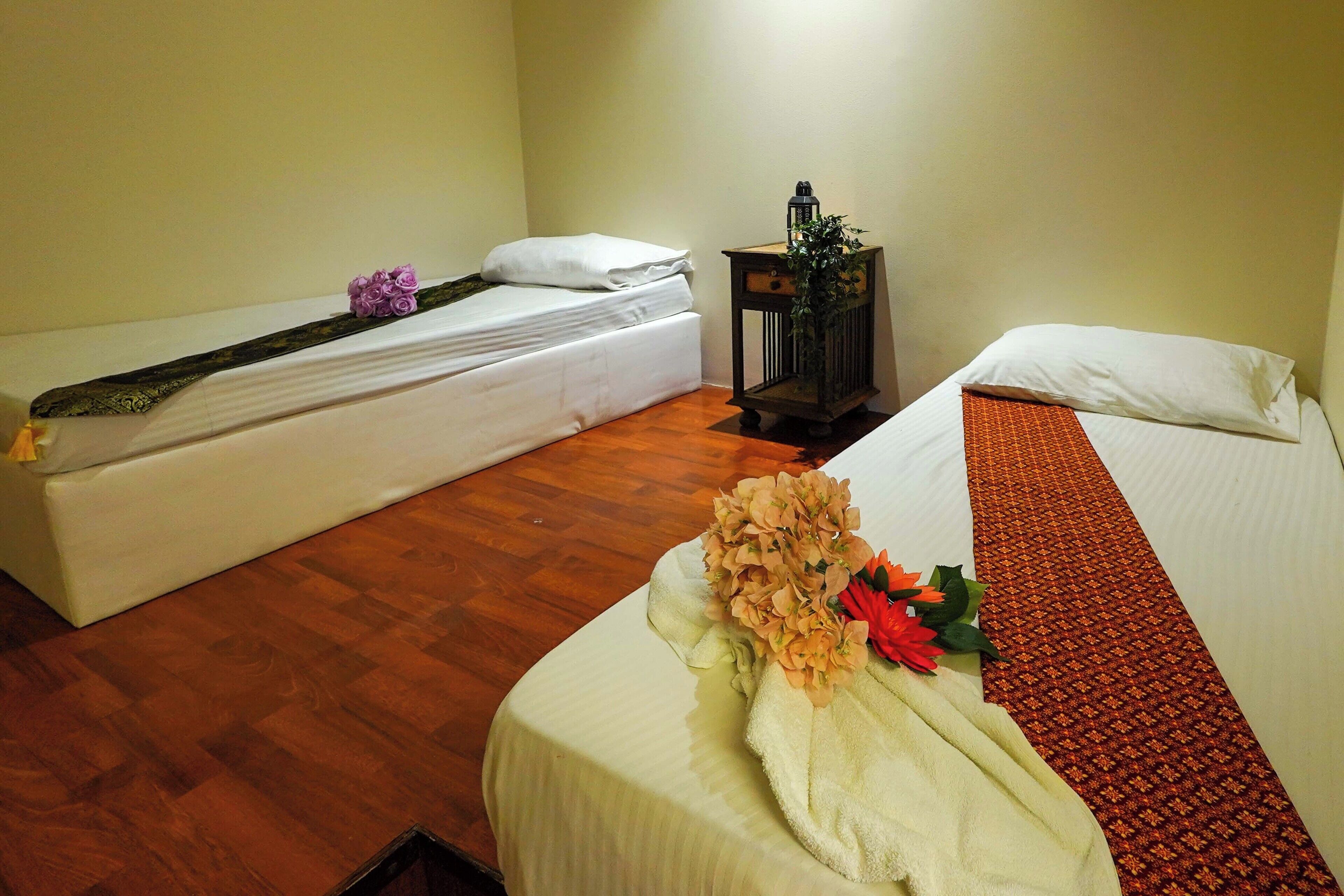 A hotel room with two beds, wooden flooring, a nightstand with a lamp, and a floral arrangement on one bed.