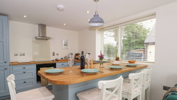 A modern kitchen with a central island, white bar stools, and a window with blinds. - Kestrel (Malpas)