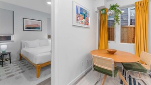 Moderni hotellihuone, jossa on sänky, ruokapöytä tuoleineen, pieni sivupöytä ja ikkuna, jossa on verhot.