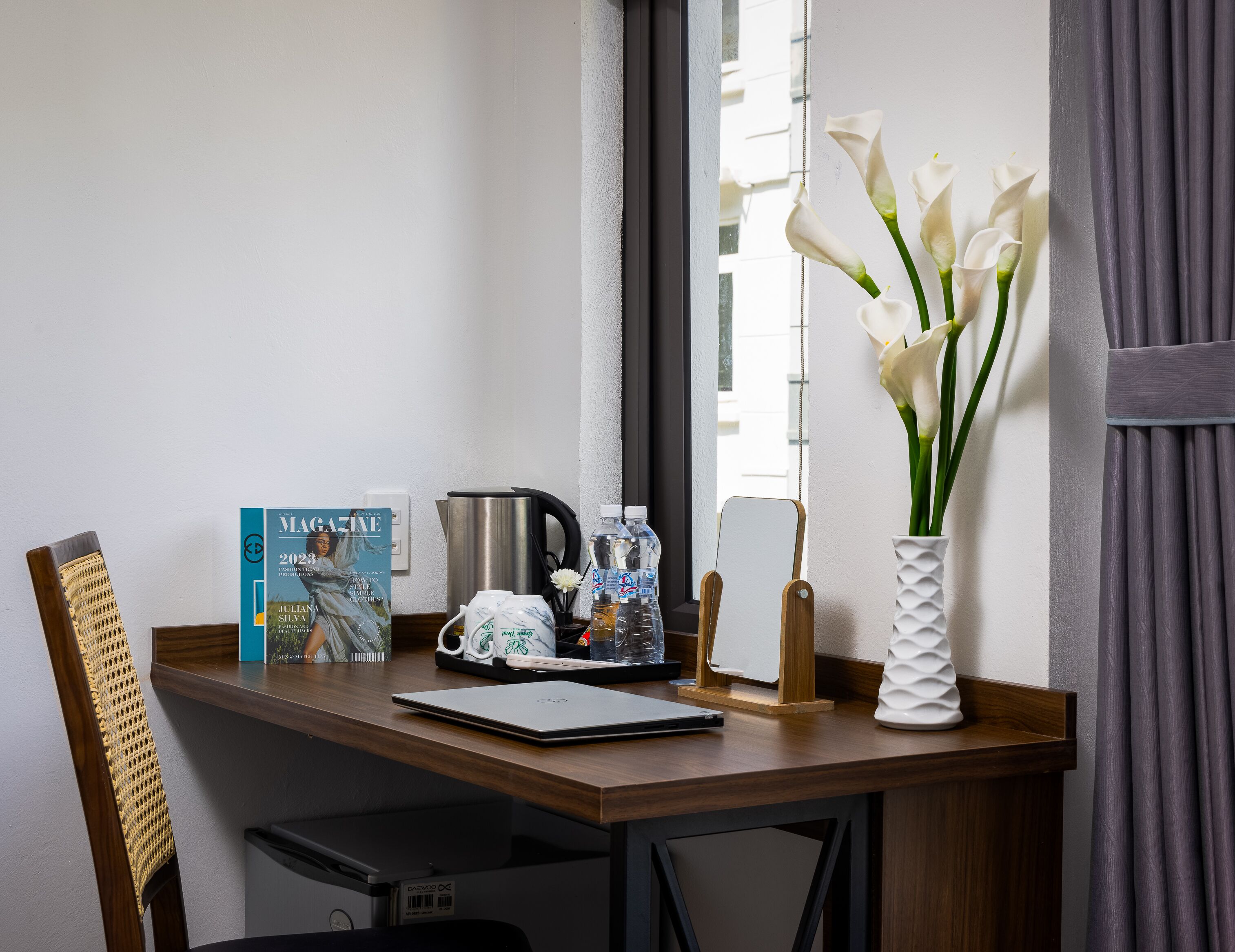 A neatly arranged desk with a laptop, a magazine, a kettle, and a vase with white flowers.