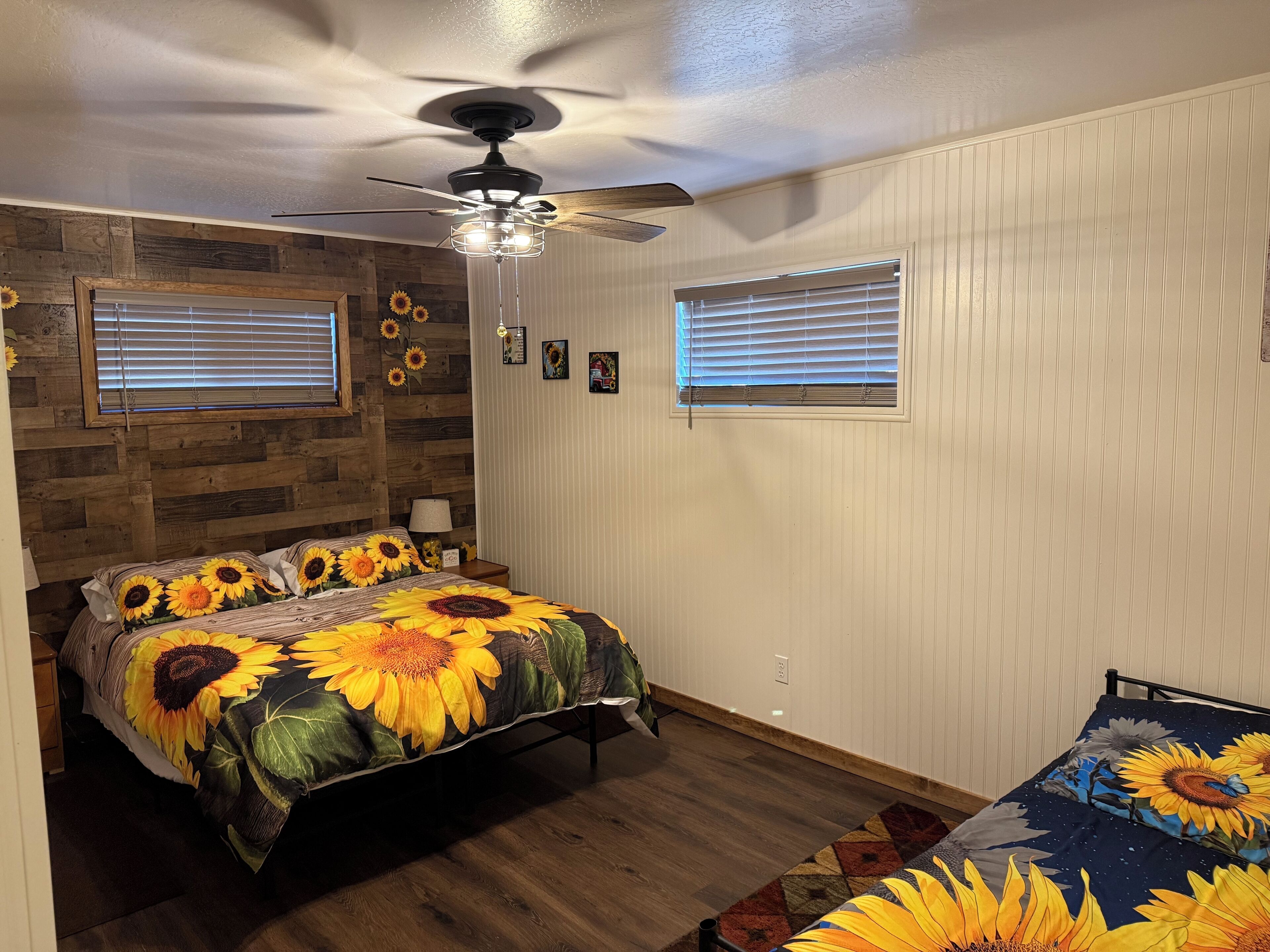 Une chambre à coucher avec un lit agrémenté d’une couvre-lit aux motifs de tournesols, un ventilateur de plafond et deux fenêtres équipées de stores.