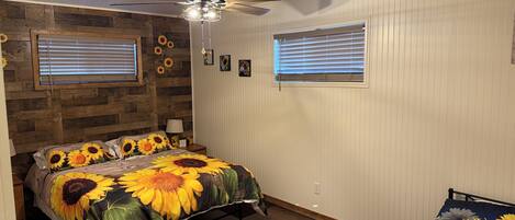 Un dormitorio con una cama con edredón con motivos de girasoles, un ventilador de techo y dos ventanas con persianas.