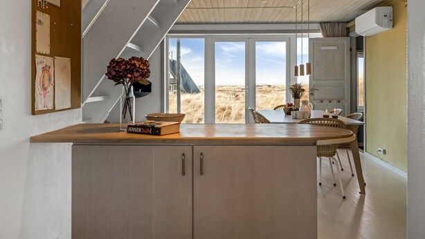 Eine moderne Küche mit einer Holzinsel, Blick auf die Dünen und einem Essbereich mit Tisch und Stühlen.