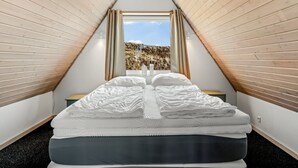 Ein Schlafzimmer mit schrägen Wänden, einem Bett mit weißer Bettwäsche und Blick auf eine grüne Landschaft durch ein Fenster.