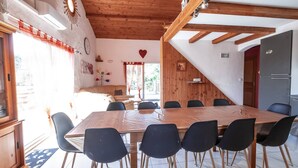 Een eetgedeelte met een grote houten tafel en zwarte stoelen, een keuken met een koelkast, en een trap die naar een ander niveau leidt.