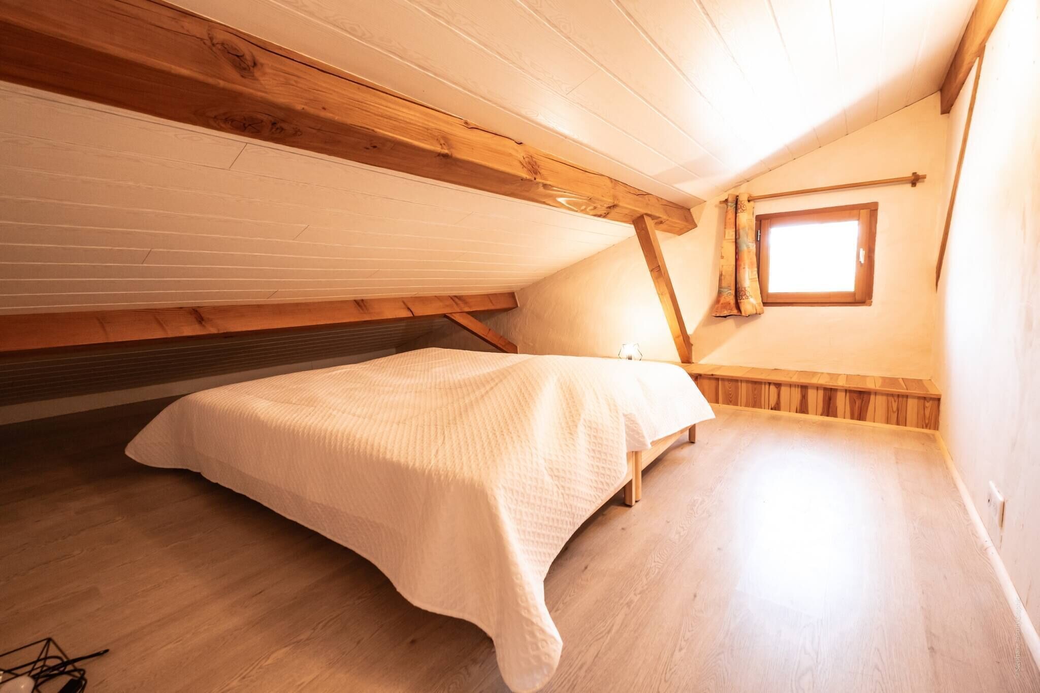 Une petite chambre mansardée, chaleureuse et accueillante, avec un plafond en bois, un lit simple et une fenêtre laissant entrer la lumière naturelle.