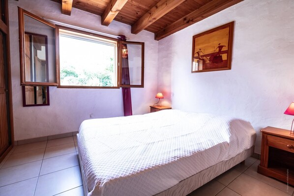 Un dormitorio con cama, ventana con cortinas, mesita de noche y un cuadro en la pared.