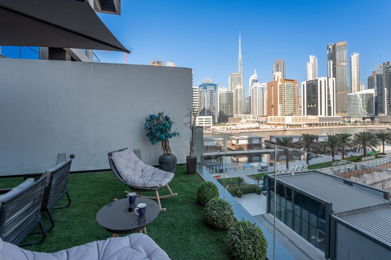 A rooftop terrace with a view of a city skyline, including a prominent skyscraper. There is a chair, a small table, and a potted plant.