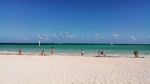 Una playa con gente caminando y nadando, cielo despejado y mar tranquilo.