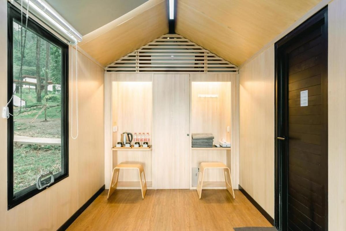 A compact room with a sloped ceiling, two wooden stools, a small table with a kettle, and a window with a view of greenery.