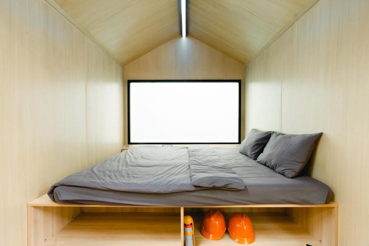 Habitación pequeña con una cama, un ventanal grande y una estantería con cascos.