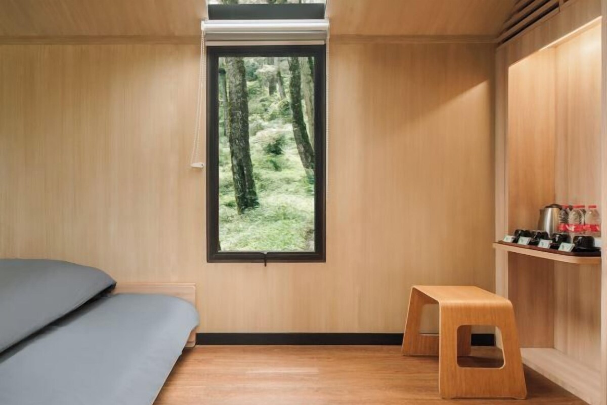 Une pièce avec un lit, une fenêtre donnant sur des arbres, un tabouret en bois et une étagère avec divers objets.