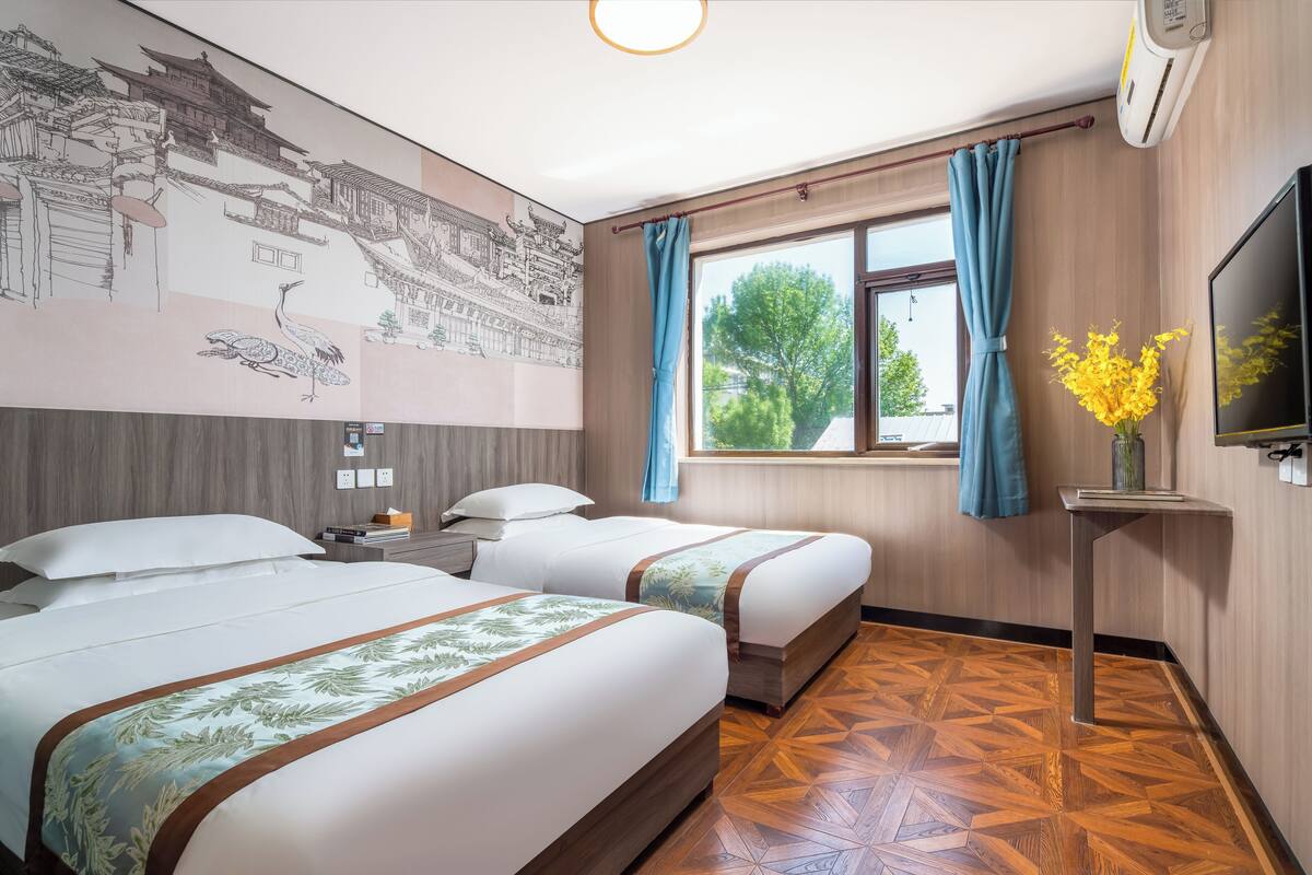 Une chambre d’hôtel avec deux lits, une grande fresque murale, une fenêtre avec des rideaux, un plancher en bois et un téléviseur fixé au mur.