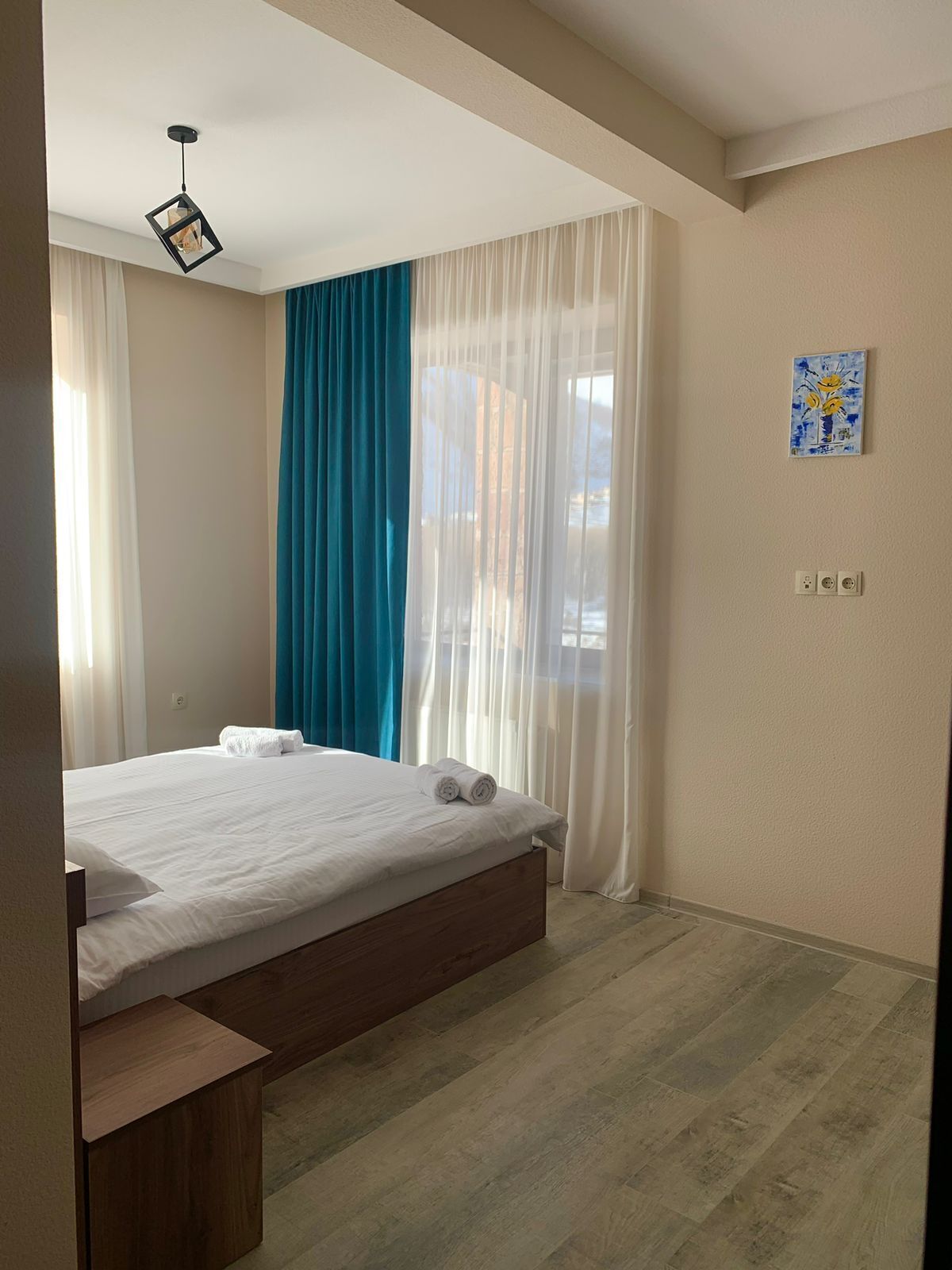 A neatly arranged bedroom with a bed, a bedside table, a window with sheer curtains, and a painting on the wall.