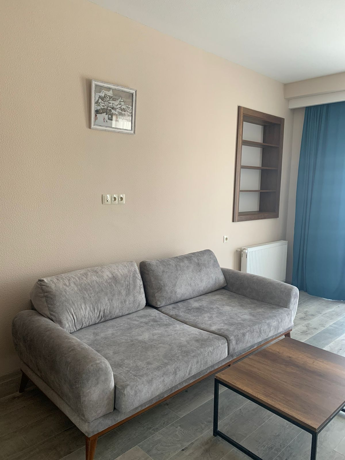A grey sofa with wooden legs, a small wooden coffee table, a wall-mounted picture, a window with a wooden frame, and a radiator.