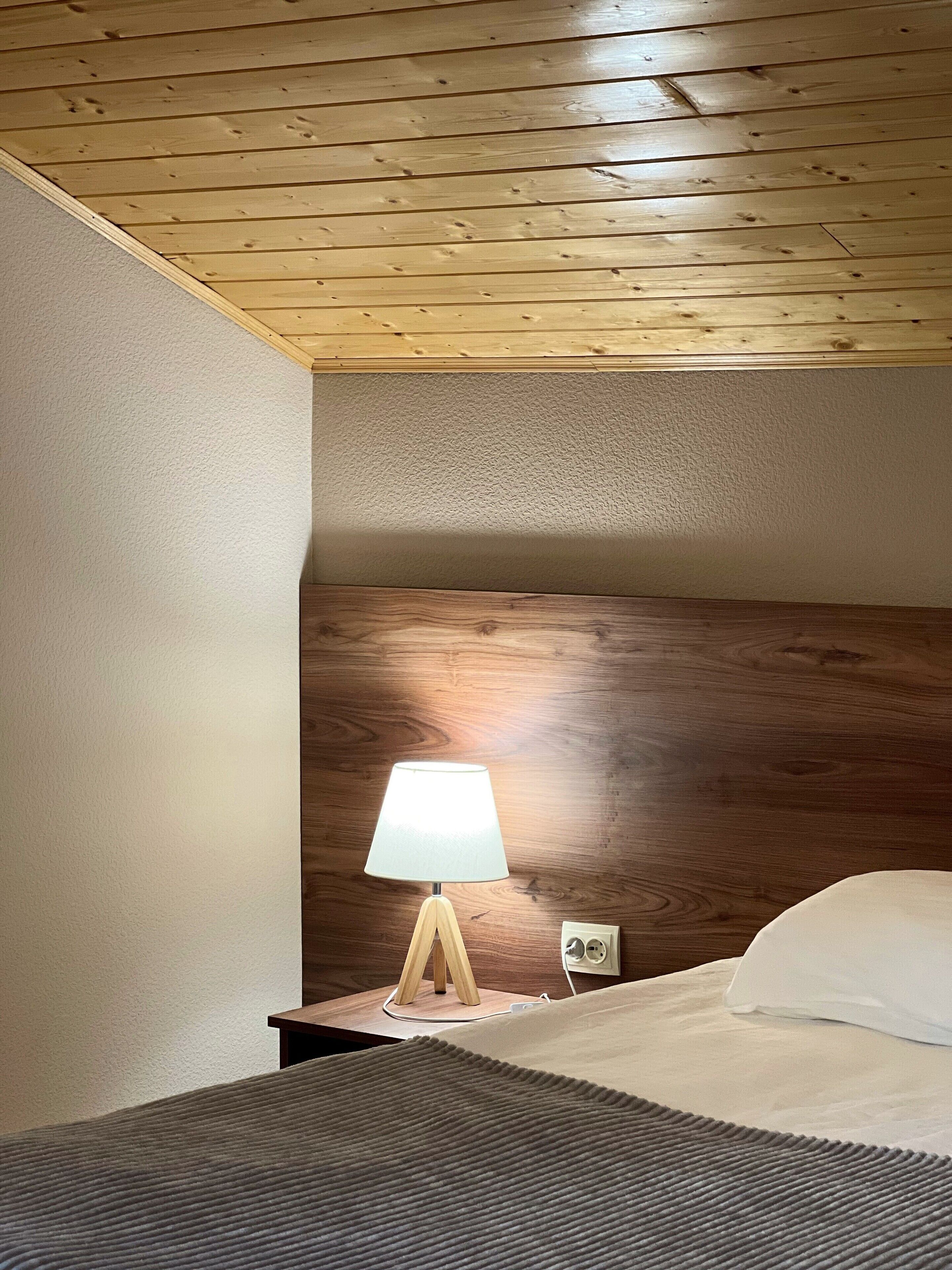 A hotel room with a wooden ceiling, a bedside lamp, a bed with white bedding, and a textured rug.