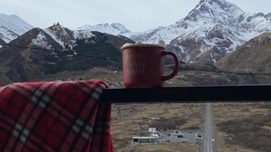 Una taza roja sobre un alféizar con un paño escocés, con vistas a una carretera y montañas nevadas.