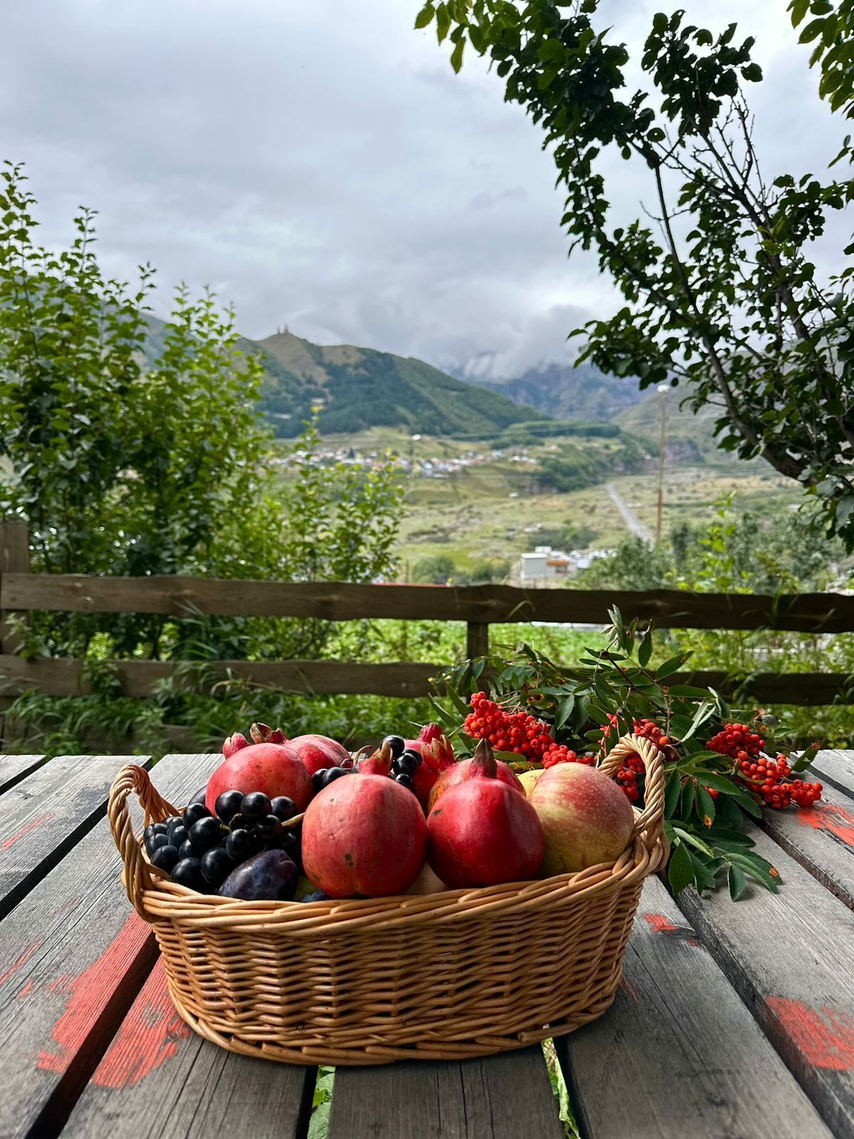 A wicker basket filled with fruits on a wooden table with a scenic mountain view in the background.