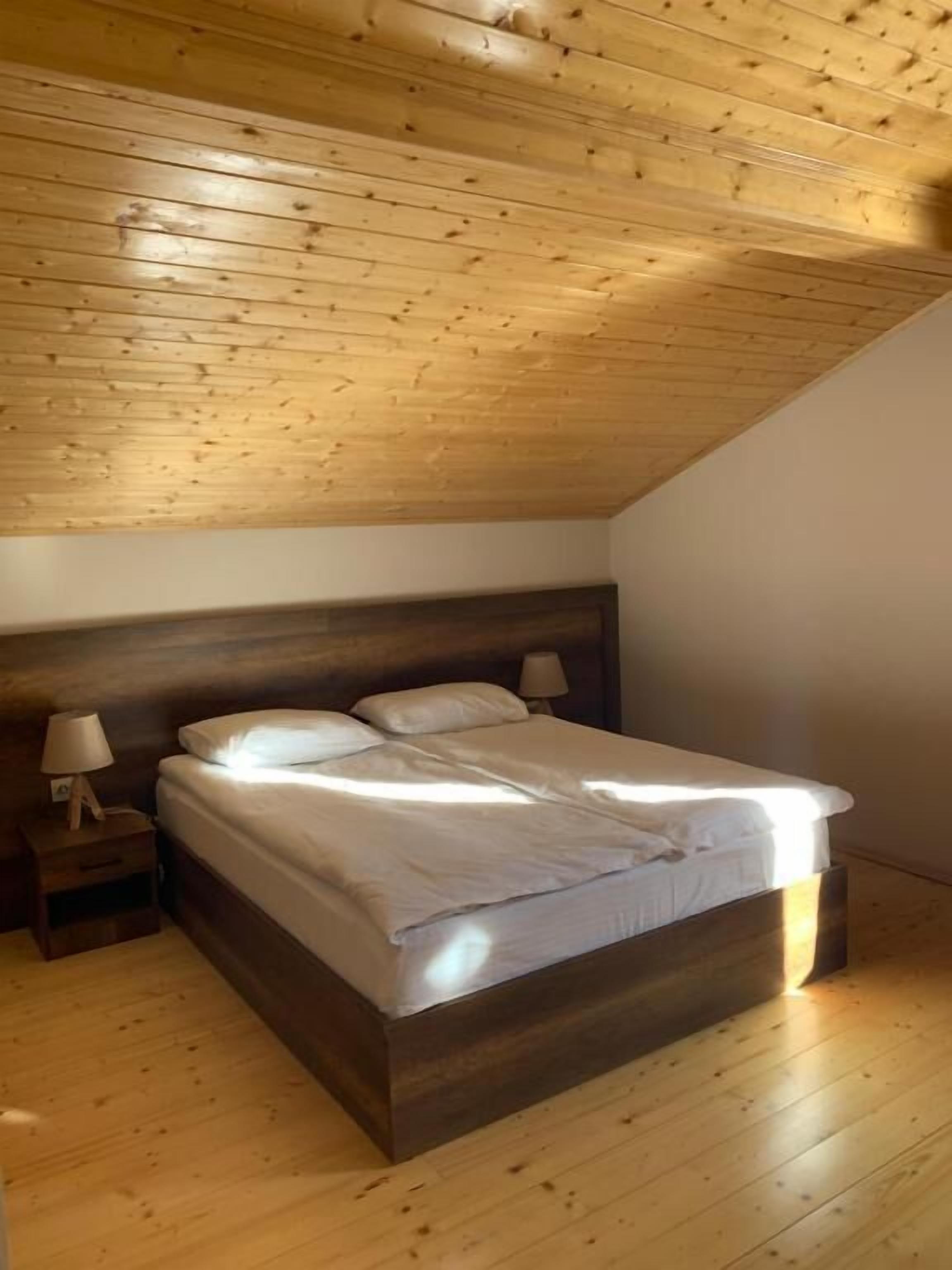 A bedroom with a wooden ceiling, a bed with white linens, two bedside tables with lamps, and a wooden floor.
